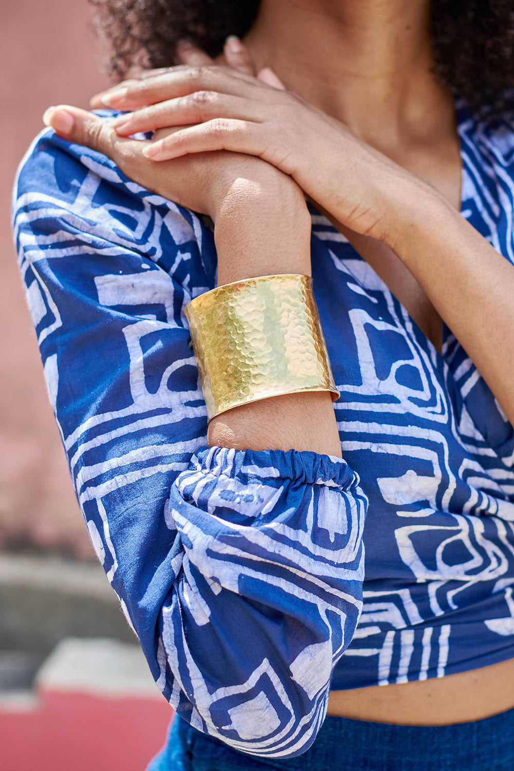 Close-up of woman wearing blue hand-dyed batik Coco print long sleeve wrap top by GEOMETRIC and brass bracelet. 