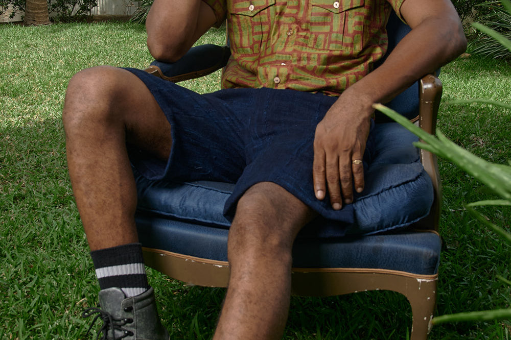 Man sitting in chair wearing hand-dyed green batik Garden Gate print short sleeve button up shirt by GEOMETRIC. 