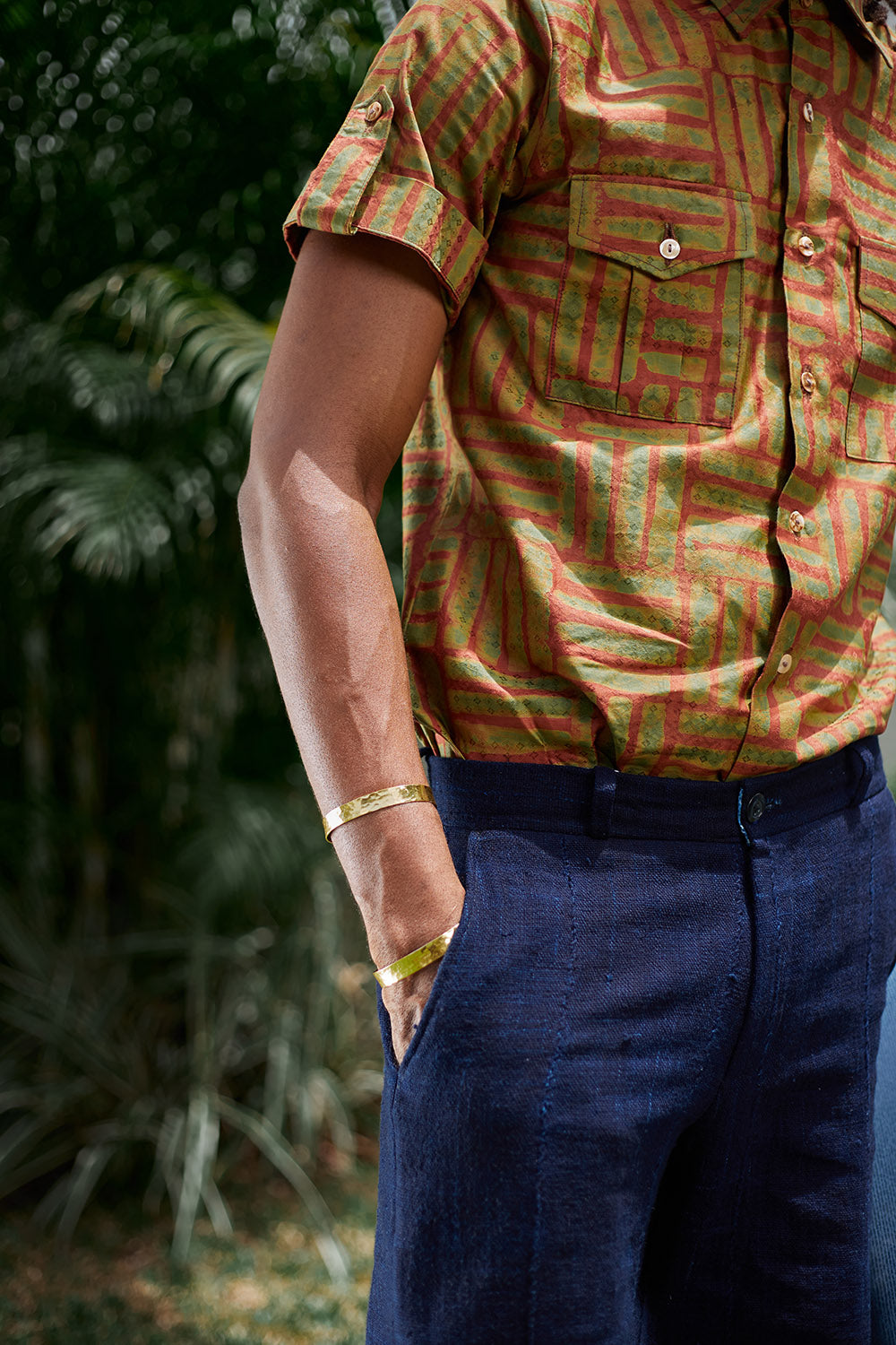 Man wearing blue shorts and hand-dyed green batik Garden Gate print short sleeve button up shirt by GEOMETRIC. 